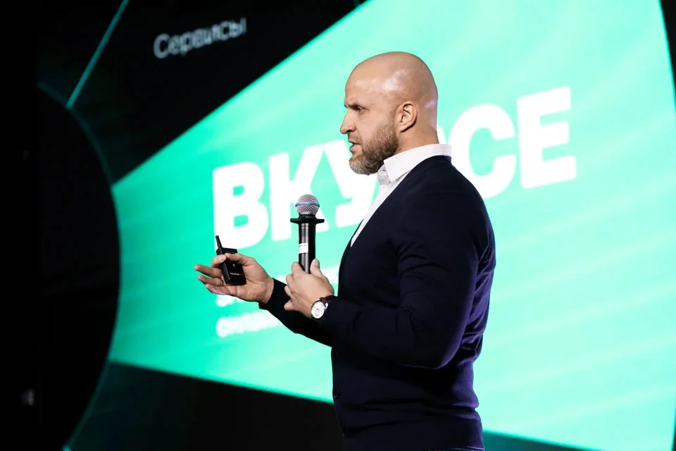 Юрий Матвеев выступает на IVA DAY 2024 на фоне яркого экрана, с микрофоном и кликером